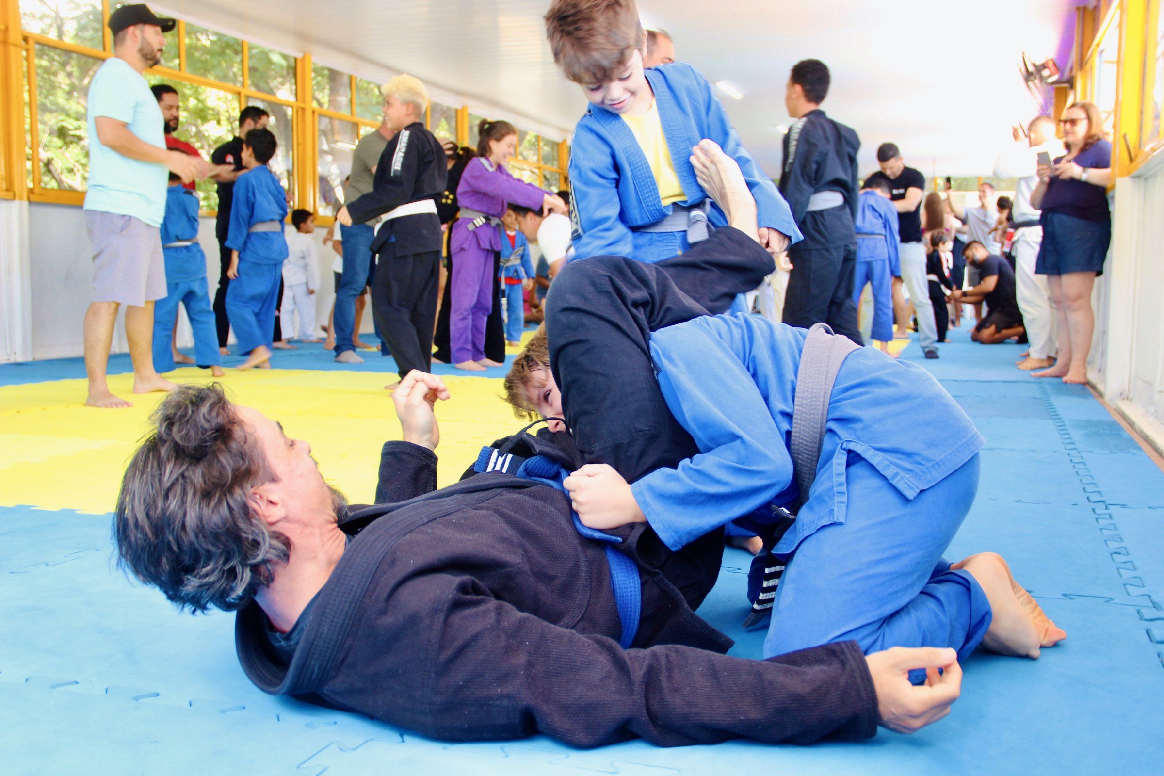 Aluno adulto e criança treinando jiu-jitsu no tatame, com famílias assistindo ao fundo