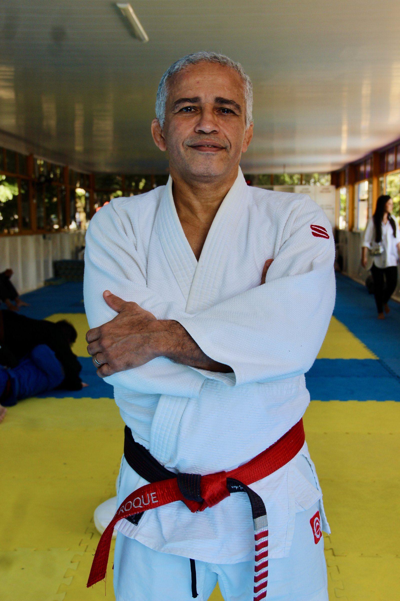 Mestre João Roque de quimono branco e faixa vermelha, de braços cruzados no tatame